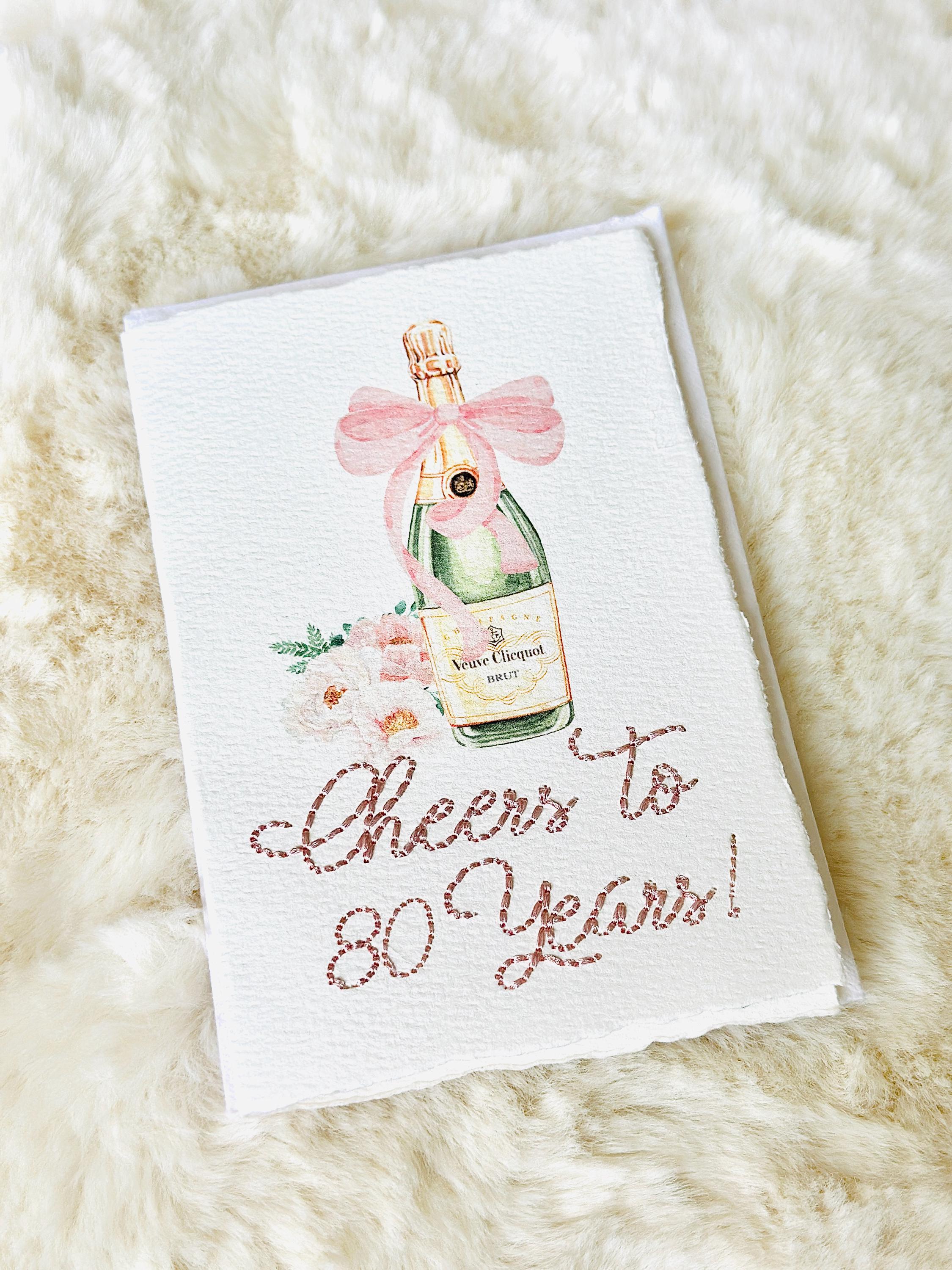 A birthday card with a watercolor champagne bottle with a pink bow and flowers to the left side. Under the art is an embroidered message: Cheers to 80 Years in pink thread. The card and matching envelope are set on a fur blanket.