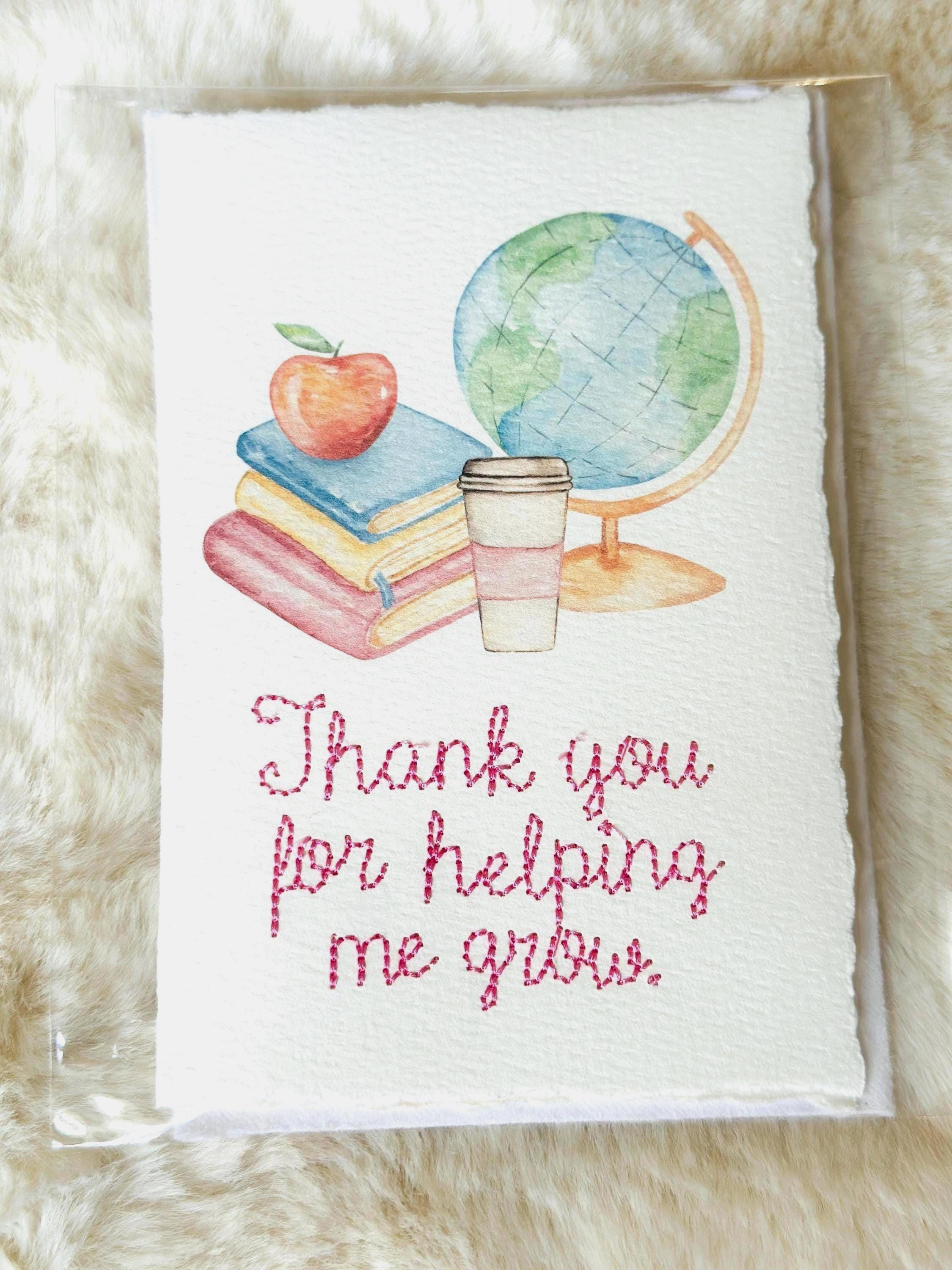 A handmade card that has watercolor art of a pile of books with an apple on top next to a globe and a to-go cup of coffee in soft pastel colors with embroidery under it that says: Thank you for helping me grow. The embroidery is done in pink thread. The card is set on a fur mat.