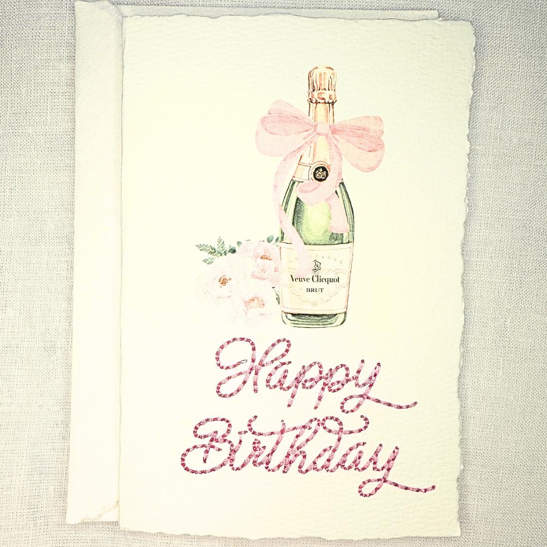 A deckled edge card with watercolor art of a champagne bottle with a pink bow and pink flowers to the left. Under the art is embroidered: Happy Birthday in pink. The card is set on a white linen table. Cards are approximately 4x6.