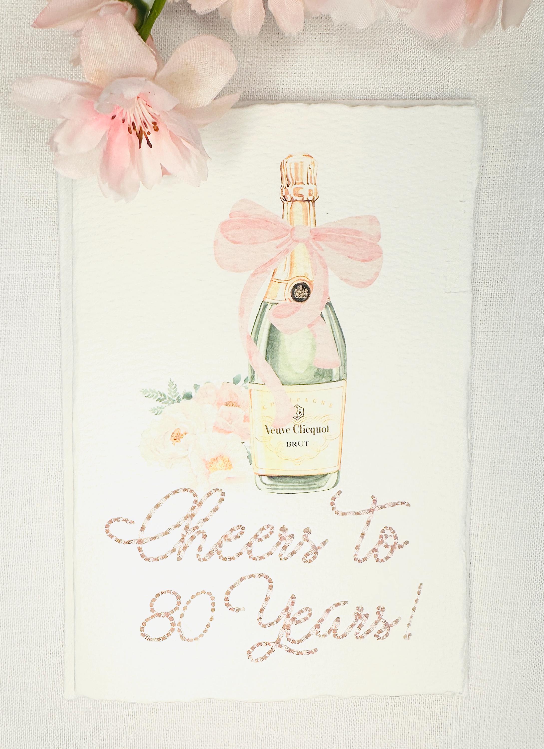 A birthday card with a watercolor champagne bottle with a pink bow and flowers to the left side. Under the art is an embroidered message: Cheers to 80 Years in pink thread. The card and matching envelope are set on a white linen tablecloth with pink flowers on the top of the card.
