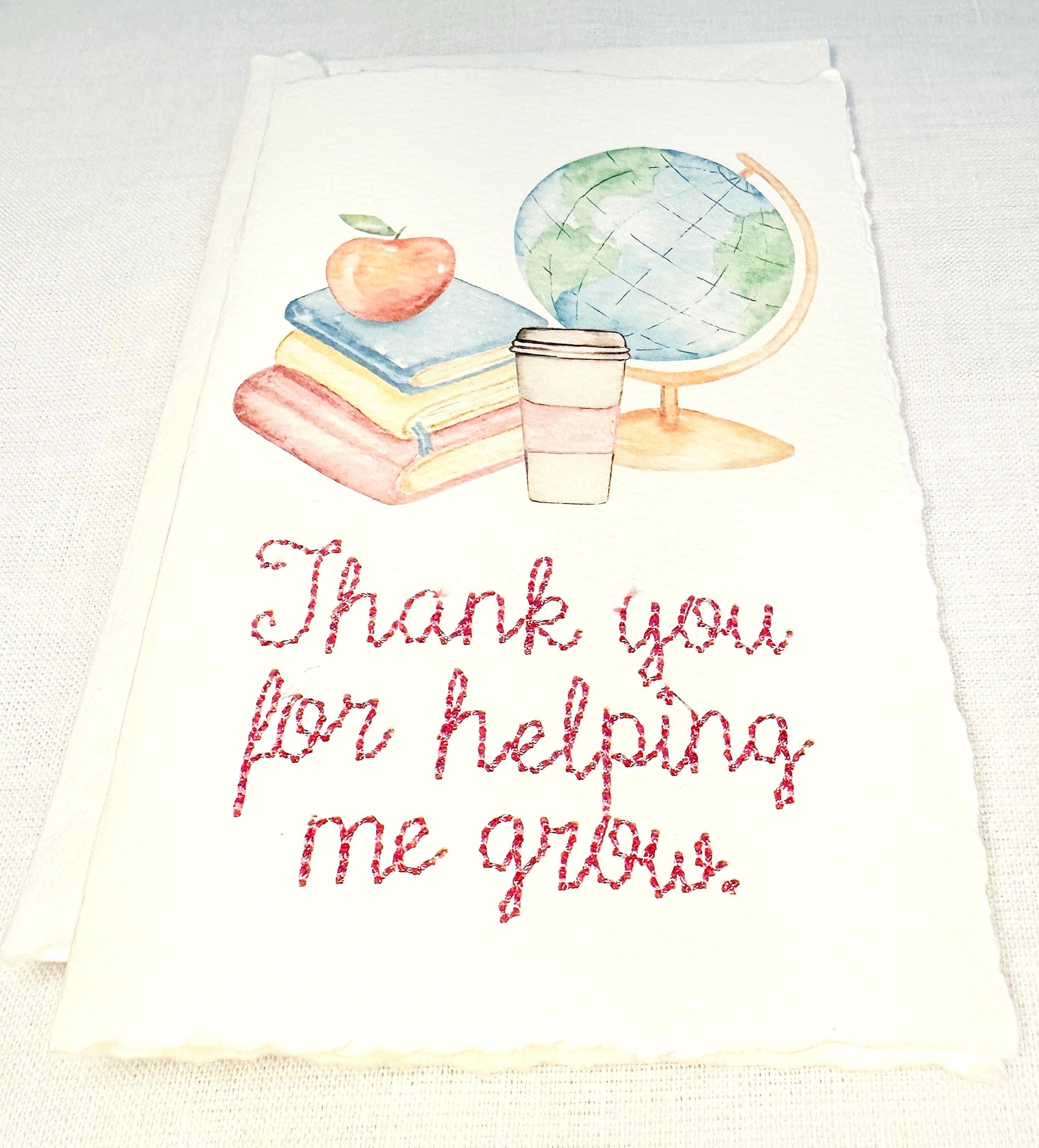 A handmade card that has watercolor art of a pile of books with an apple on top next to a globe and a to-go cup of coffee in soft pastel colors with embroidery under it that says: Thank you for helping me grow. The embroidery is done in pink thread. The card is set on a white table.