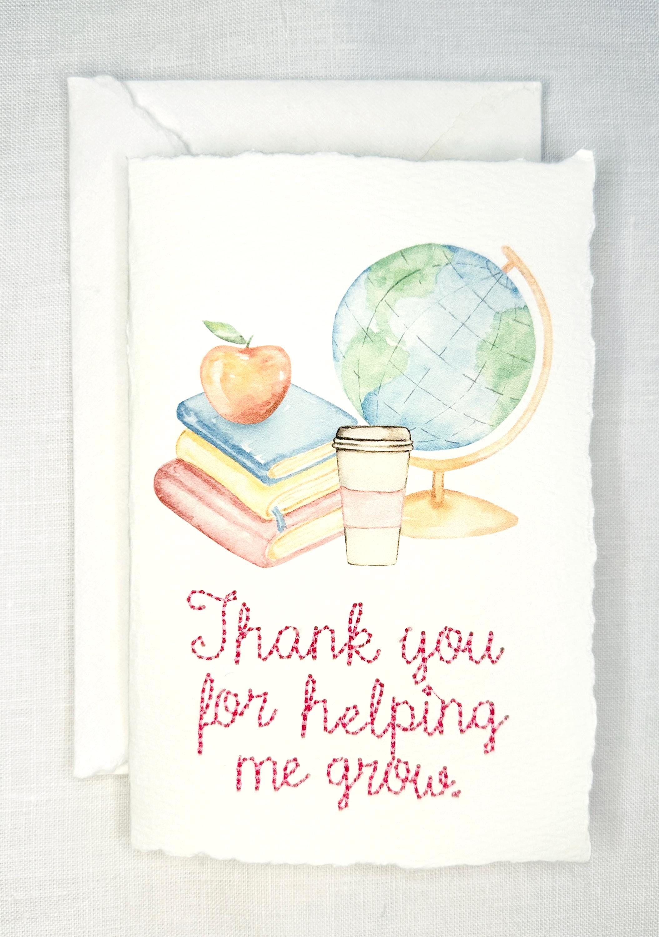 A handmade card that has watercolor art of a pile of books with an apple on top next to a globe and a to-go cup of coffee in soft pastel colors with embroidery under it that says: Thank you for helping me grow. The embroidery is done in pink thread. The card is set on a white table.