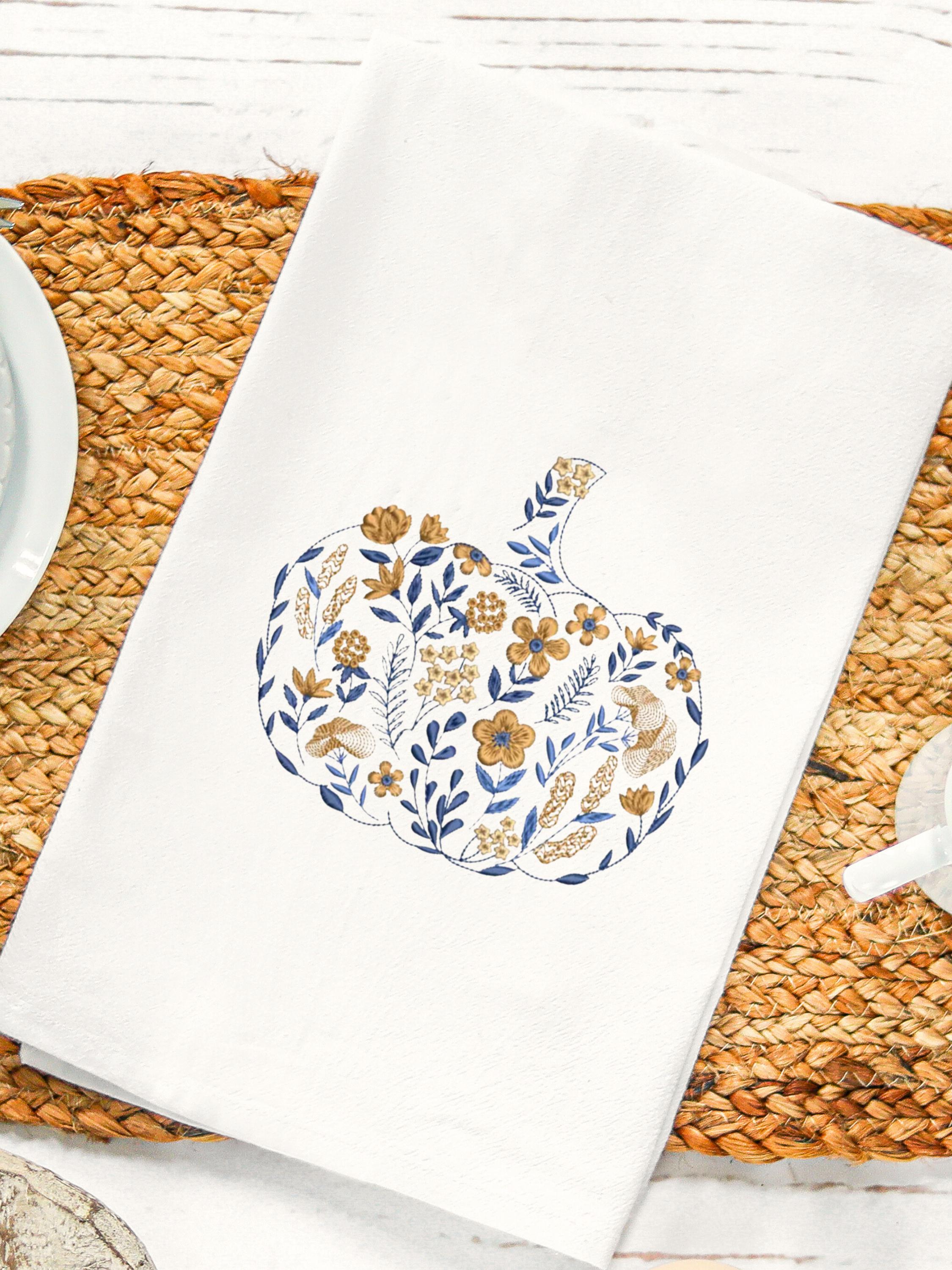 A blue and orange pumpkin that is embroidered in a chinoiserie coquette style filled with flowers of all different sizes on a white tea towel measuring 17x30" is on a straw placemat with a white saucer to the left and a white tea cup to the right.
