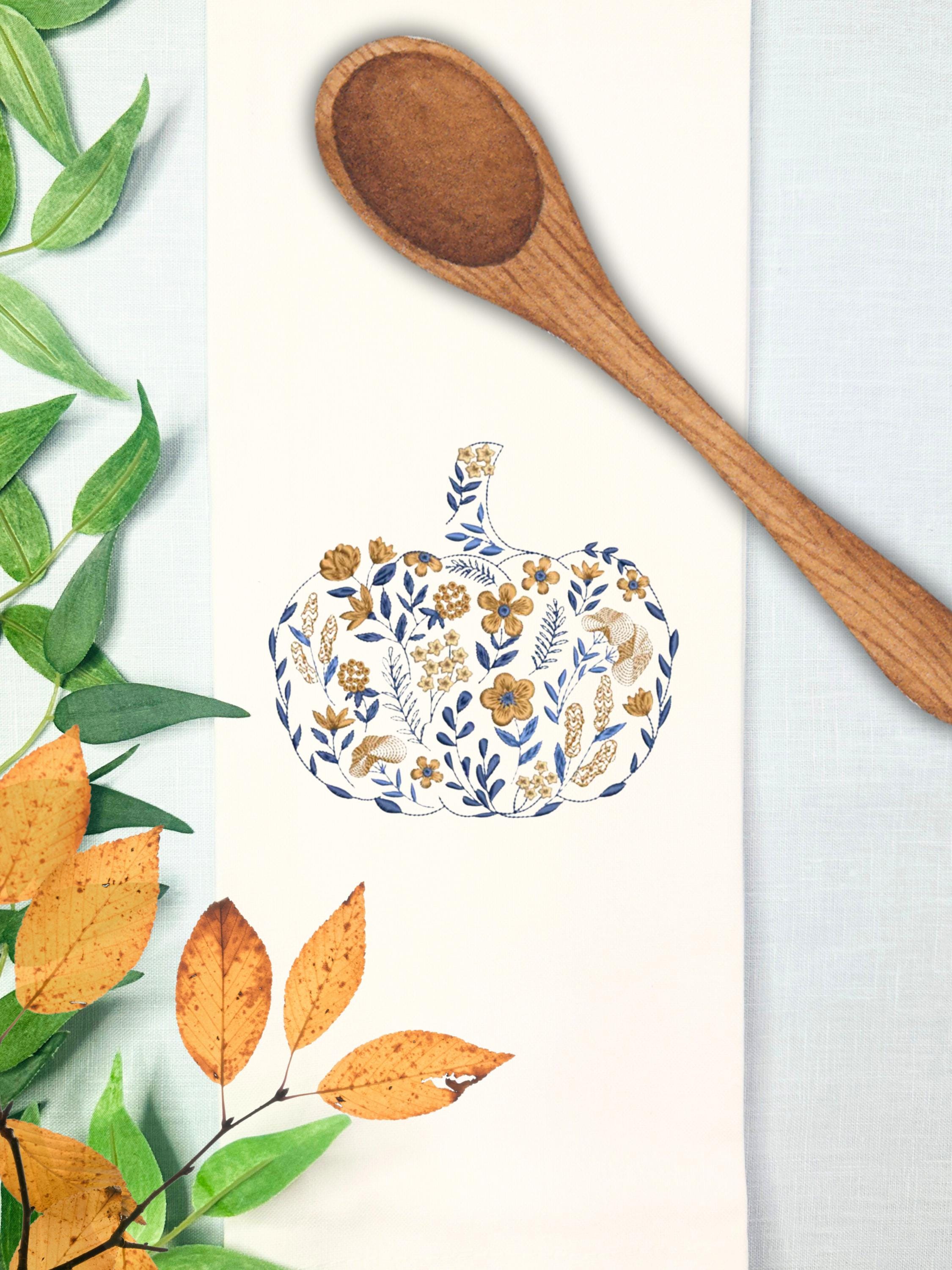 A blue and orange pumpkin that is embroidered in a chinoiserie coquette style filled with flowers of all different sizes on a white tea towel measuring 17x30" is on a light blue linen tablecloth with green and orange leaves to the left and a wooden spoon on the top.