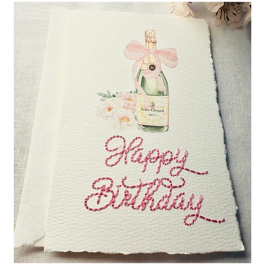 A close up of a deckled edge card with watercolor art of a champagne bottle with a pink bow and pink flowers to the left. Under the art is embroidered: Happy Birthday in pink. The card is set on a white linen table with pink cherry blossoms to the top right. Cards are approximately 4x6.