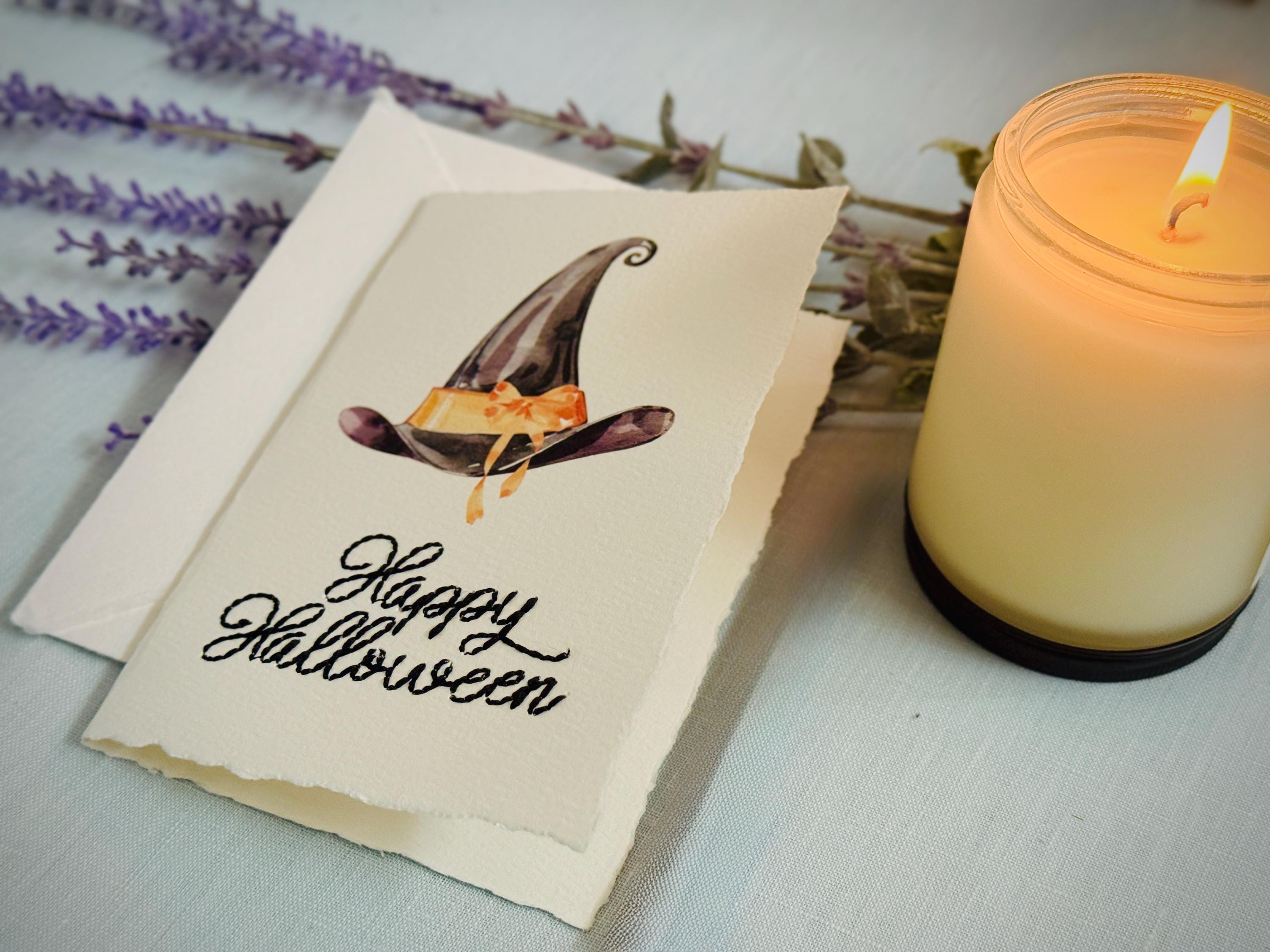 A photo of a handmade card that has a watercolor witch's hat in black with an orange bow. Under the hat the card is embroidered in black thread "Happy Halloween". The card is set on top of a light blue linen tablecloth with a lit candle next to it and a purple lavender sprig behind it.