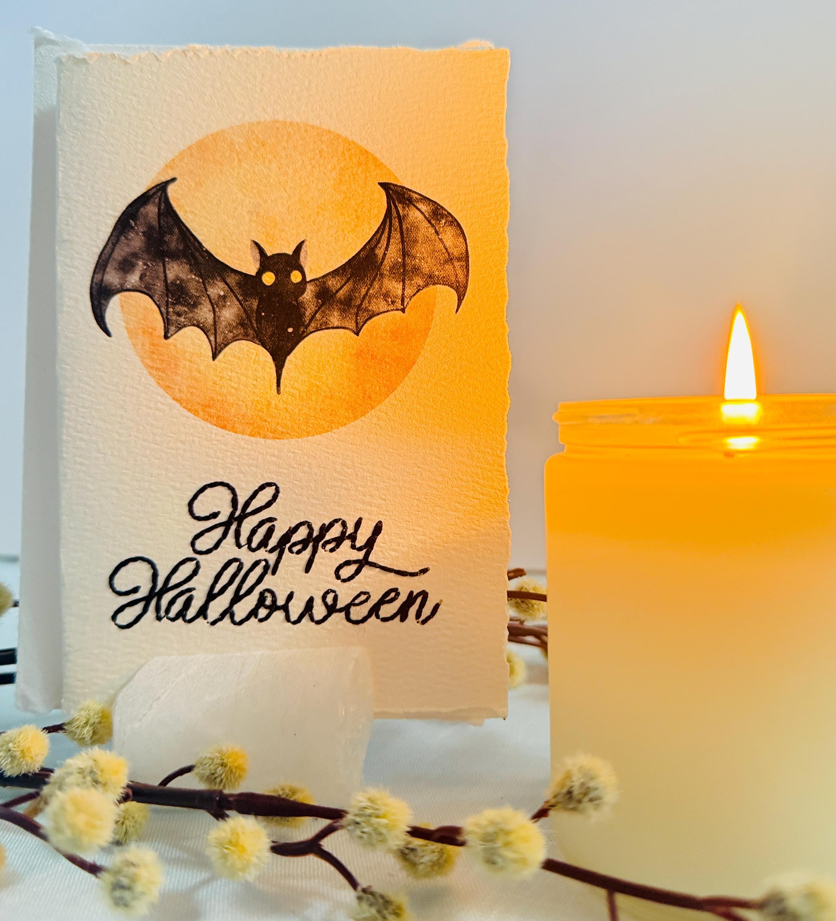 A watercolor bat in front of a moon with Happy Halloween embroidered below. The card is sitting on top of its' matching envelope in a card holder with a branch that has fuzzy white blooms on a blue linen tablecloth. Cards are approximately 4"x6". There is a lit candle to the right.