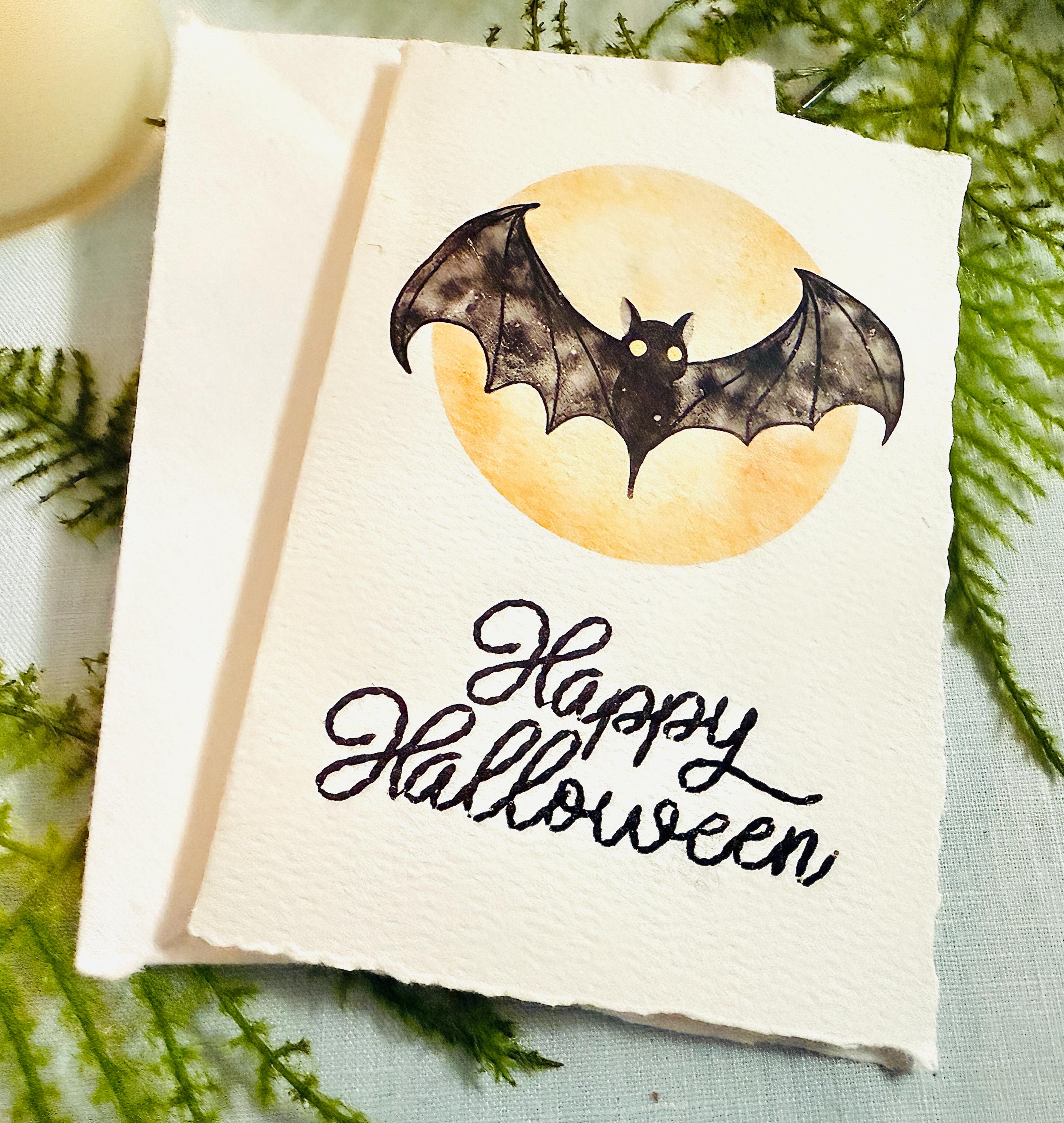 A watercolor bat in front of a moon with Happy Halloween embroidered below. The card is sitting on top of its' matching envelope which is laying on top of a fern on a blue linen tablecloth. Cards are approximately 4"x6". There is a candle to the upper left.