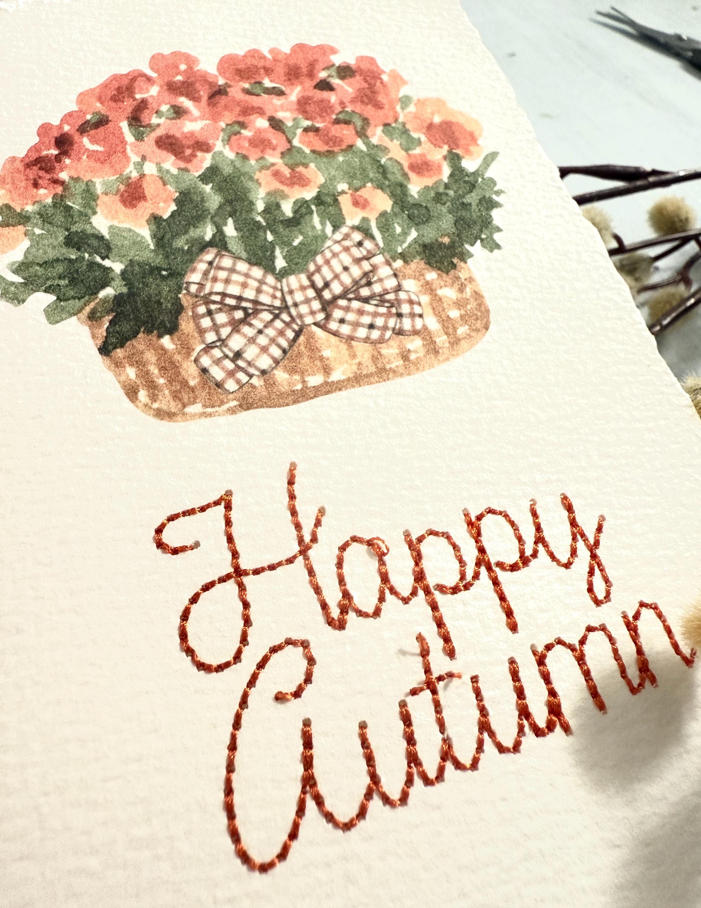 A close up of watercolor orange mums with greenery and a plaid brown bow in a mid-tone wicker basket with "Happy Autumn" embroidered below in orange.  The card is set on top of its' envelope on a blue linen tablecloth.