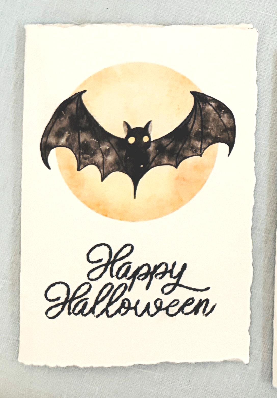A watercolor bat in front of a moon with Happy Halloween embroidered below. The card is sitting on top of its' matching envelope which is laying on top of a fern on a blue linen tablecloth. Cards are approximately 4"x6".