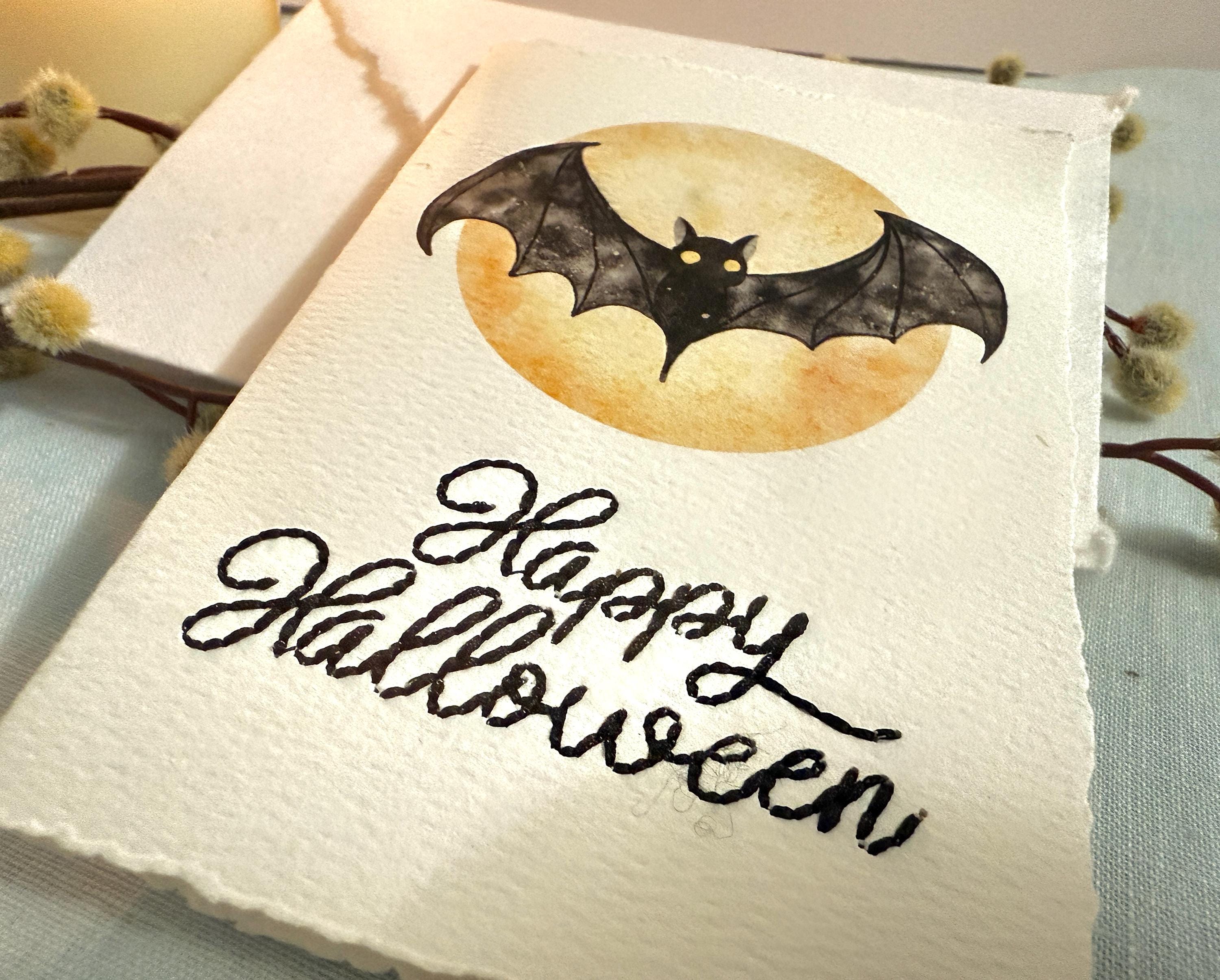 A close up of a card with a watercolor bat in front of a moon with Happy Halloween embroidered below. The card is sitting on top of its' matching envelope which is laying on top of a branch on a blue linen tablecloth. Cards are approximately 4"x6".