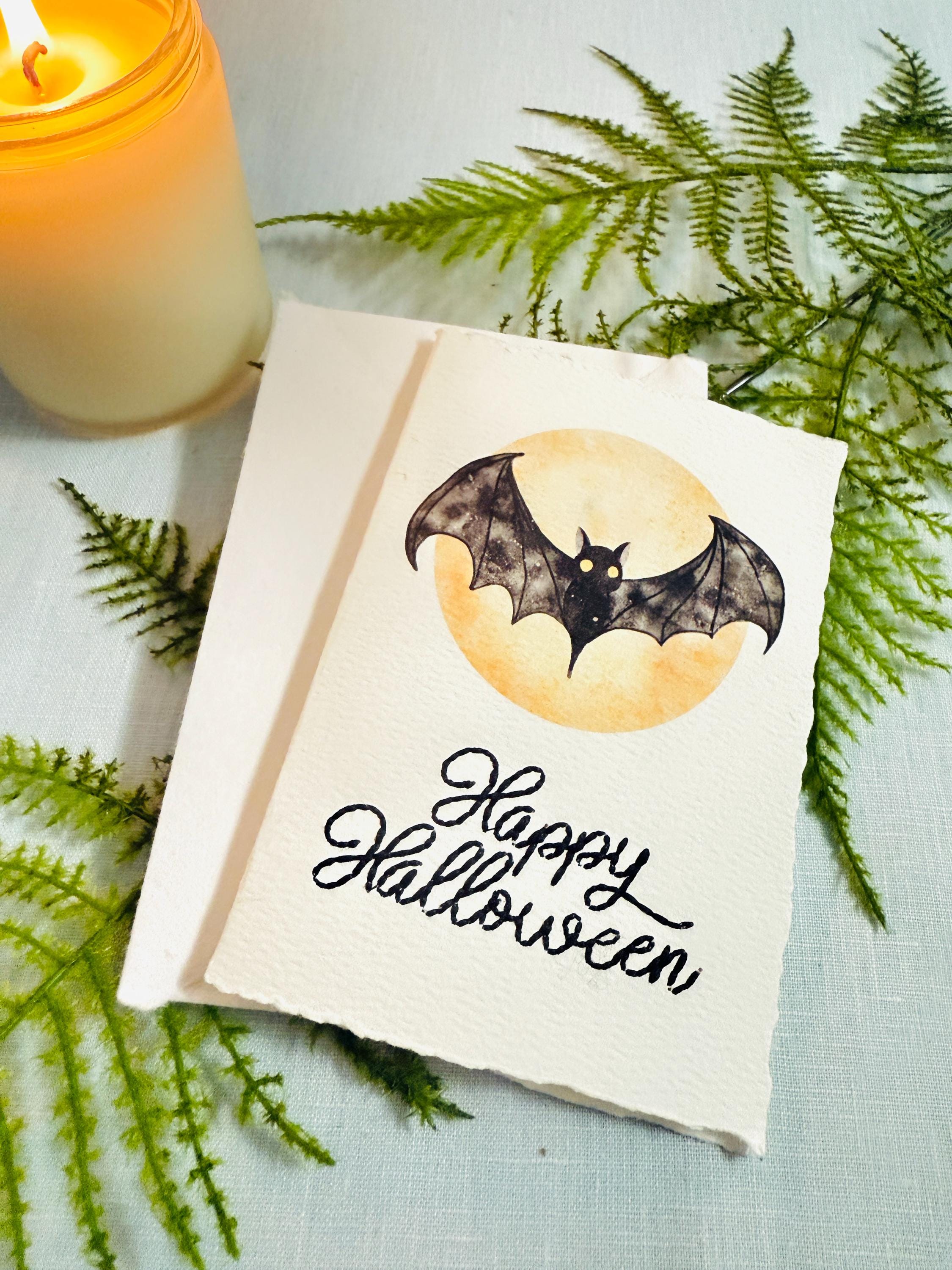 A watercolor bat in front of a moon with Happy Halloween embroidered below. The card is sitting on top of its' matching envelope which is laying on top of a fern on a blue linen tablecloth. Cards are approximately 4"x6". There is a candle to the upper right.