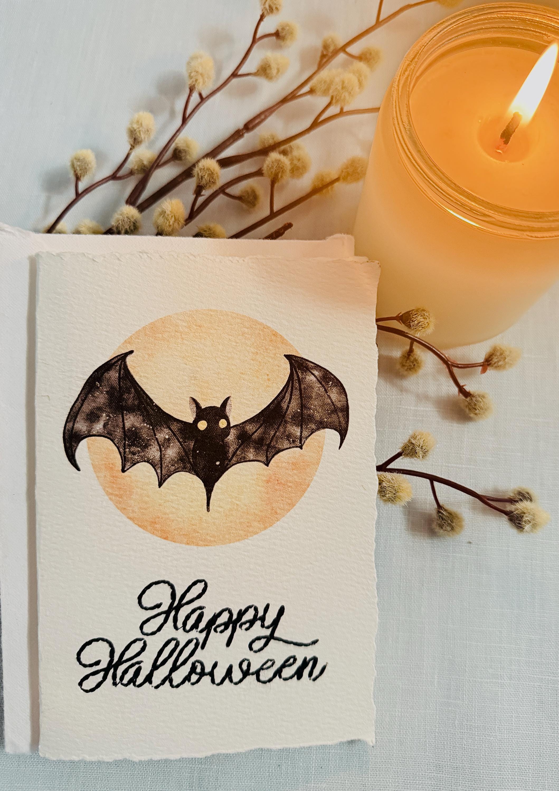 A watercolor bat in front of a moon with Happy Halloween embroidered below. The card is sitting on top of its' matching envelope which is laying on  a blue linen tablecloth. Cards are approximately 4"x6". There is a lit candle to the upper right along with a branch with fuzzy white buds.