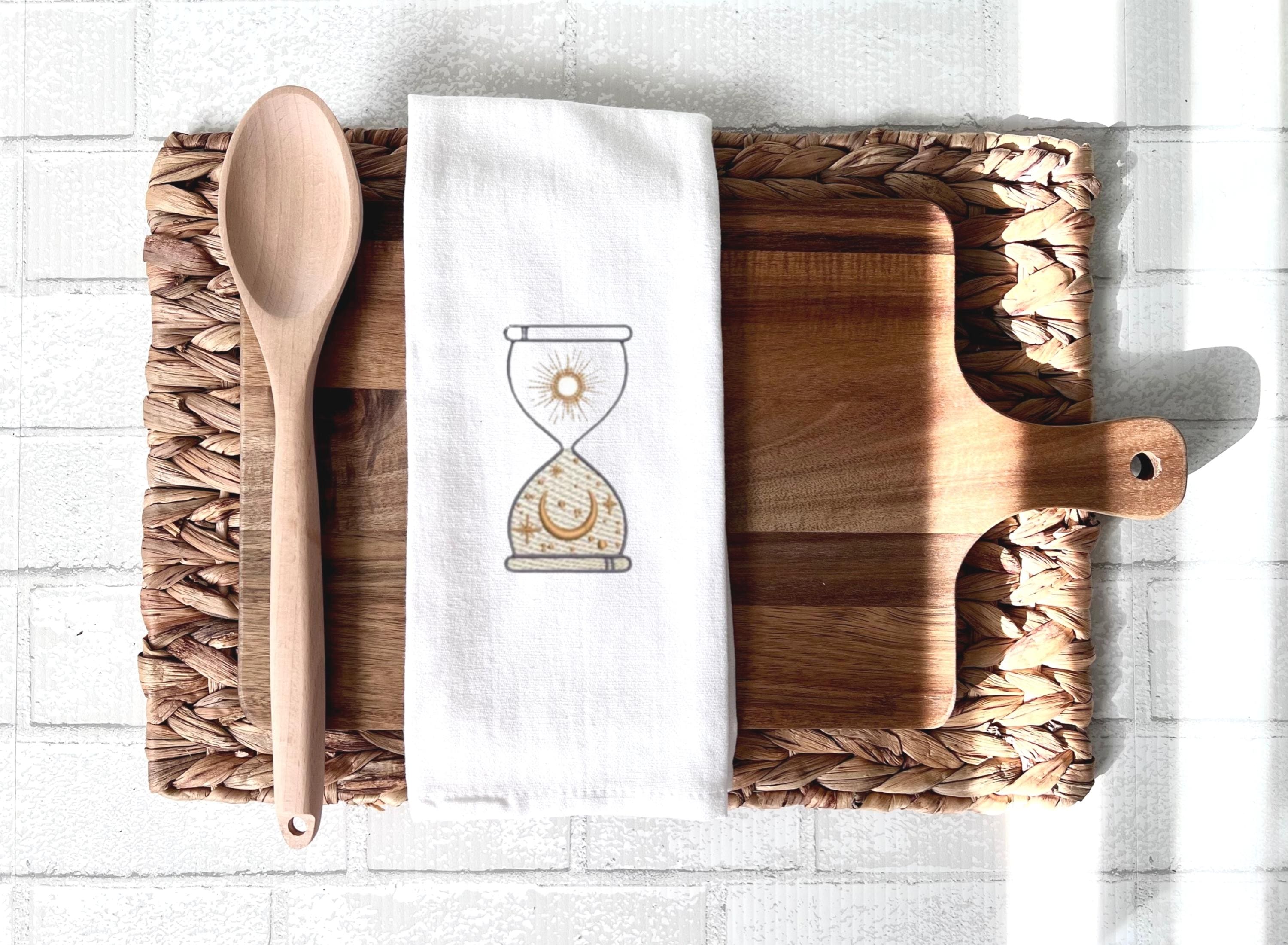 An image of an hourglass with sun and moon embroidered in gold on a white tea towel set on a cutting board with a spoon on top of a woven placemat on a white tile background.