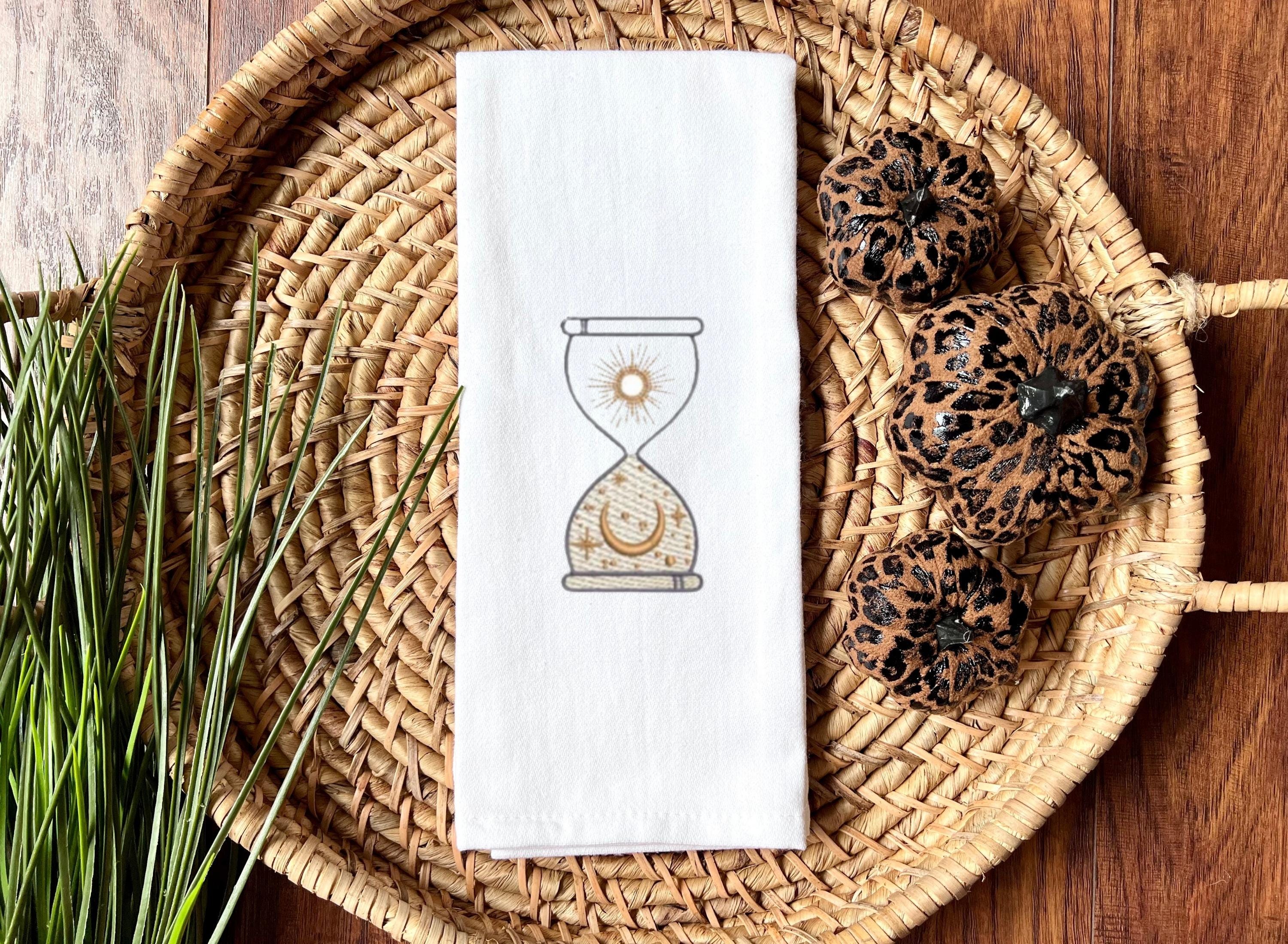 An image of an hourglass with sun and moon embroidered in gold on a white tea towel set in a basket with cheetah design pumpkins to the right and greens on the left set on a wooden table.
