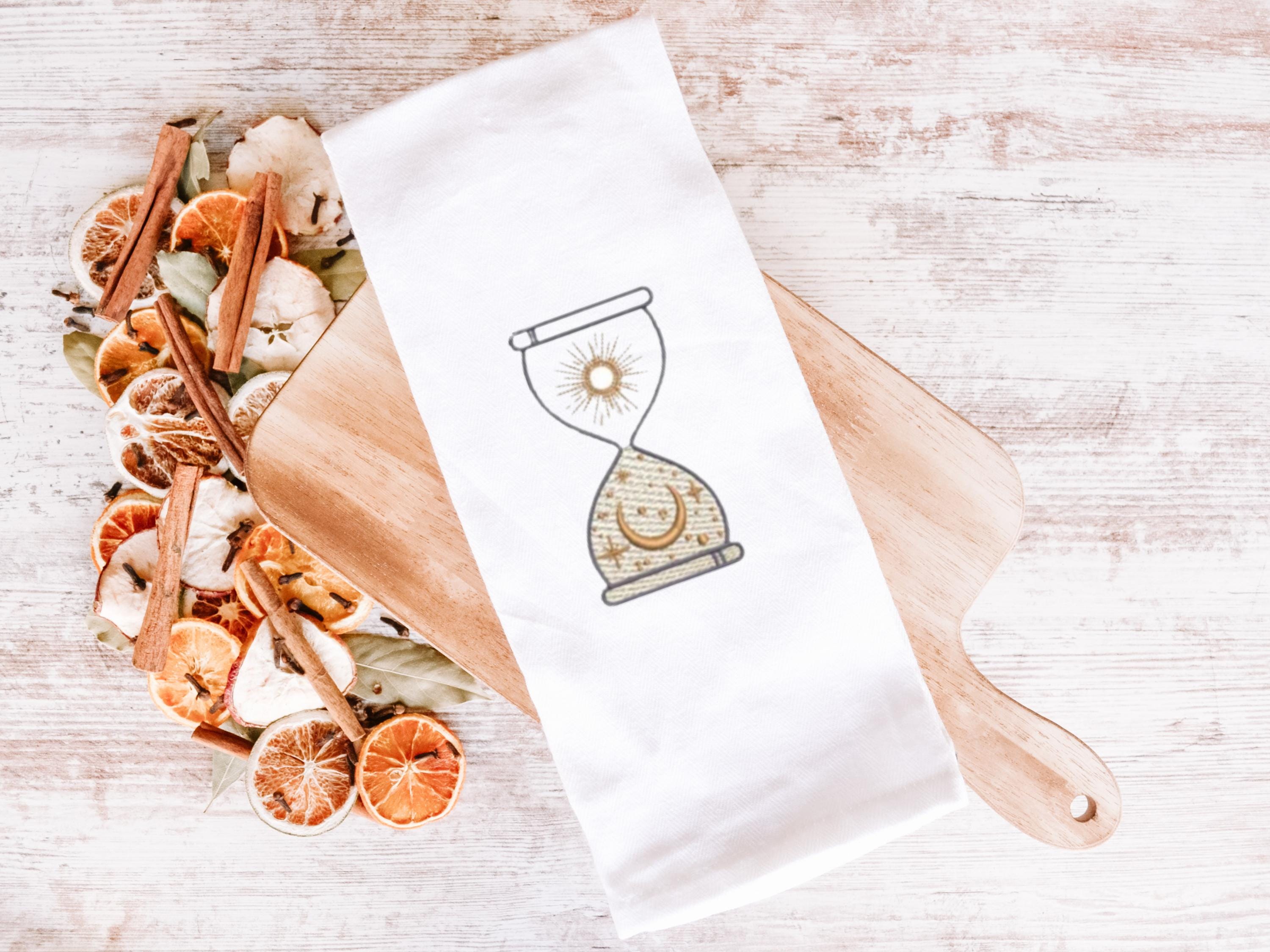 An image of an hourglass with sun and moon embroidered in gold on a white tea towel set on a wooden cutting board with dried oranges and spices to the left.
