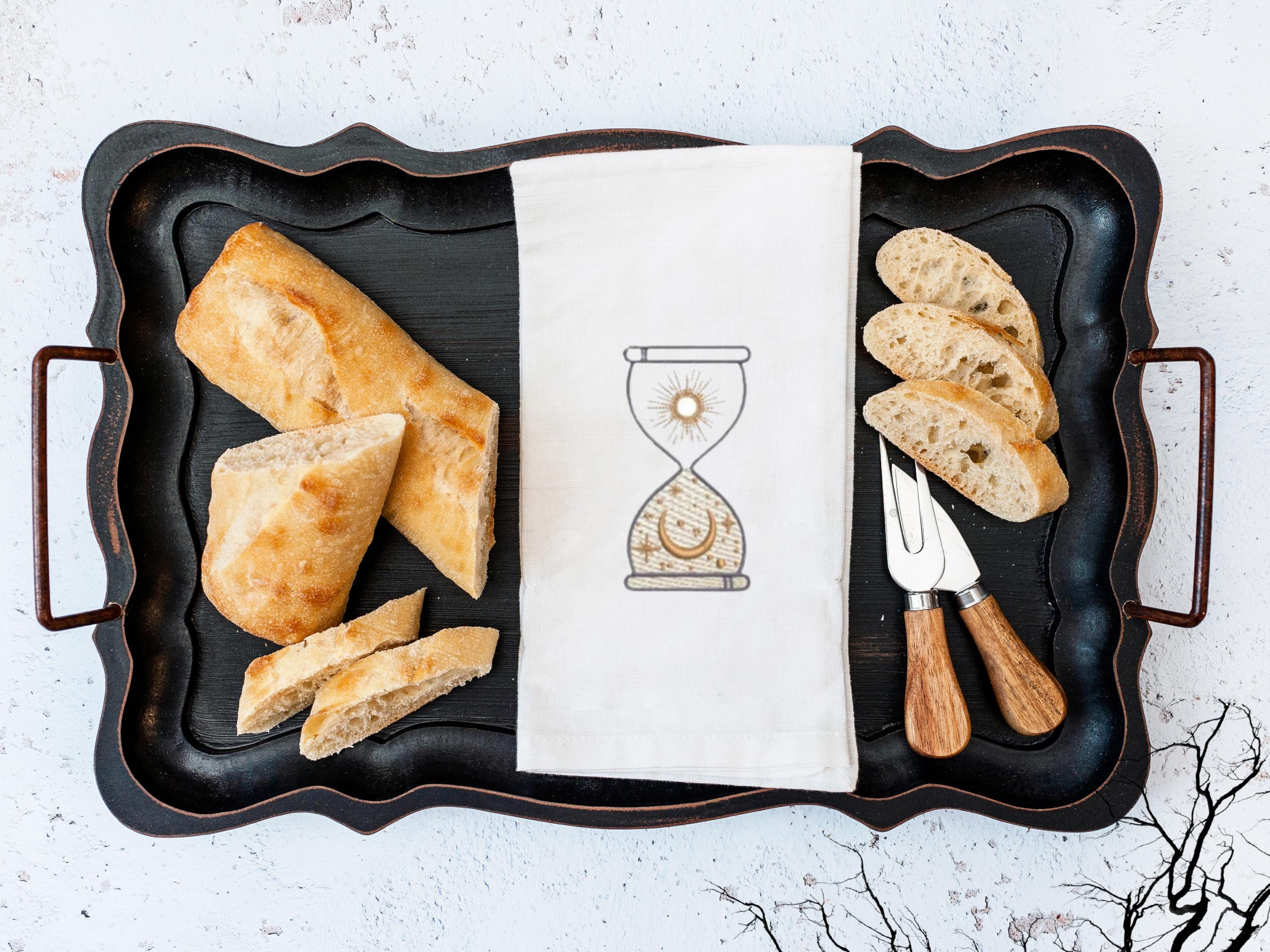 An image of an hourglass with sun and moon embroidered in gold on a white tea towel set on a black tray with bread and utensils and a few black branches to the bottom right.
