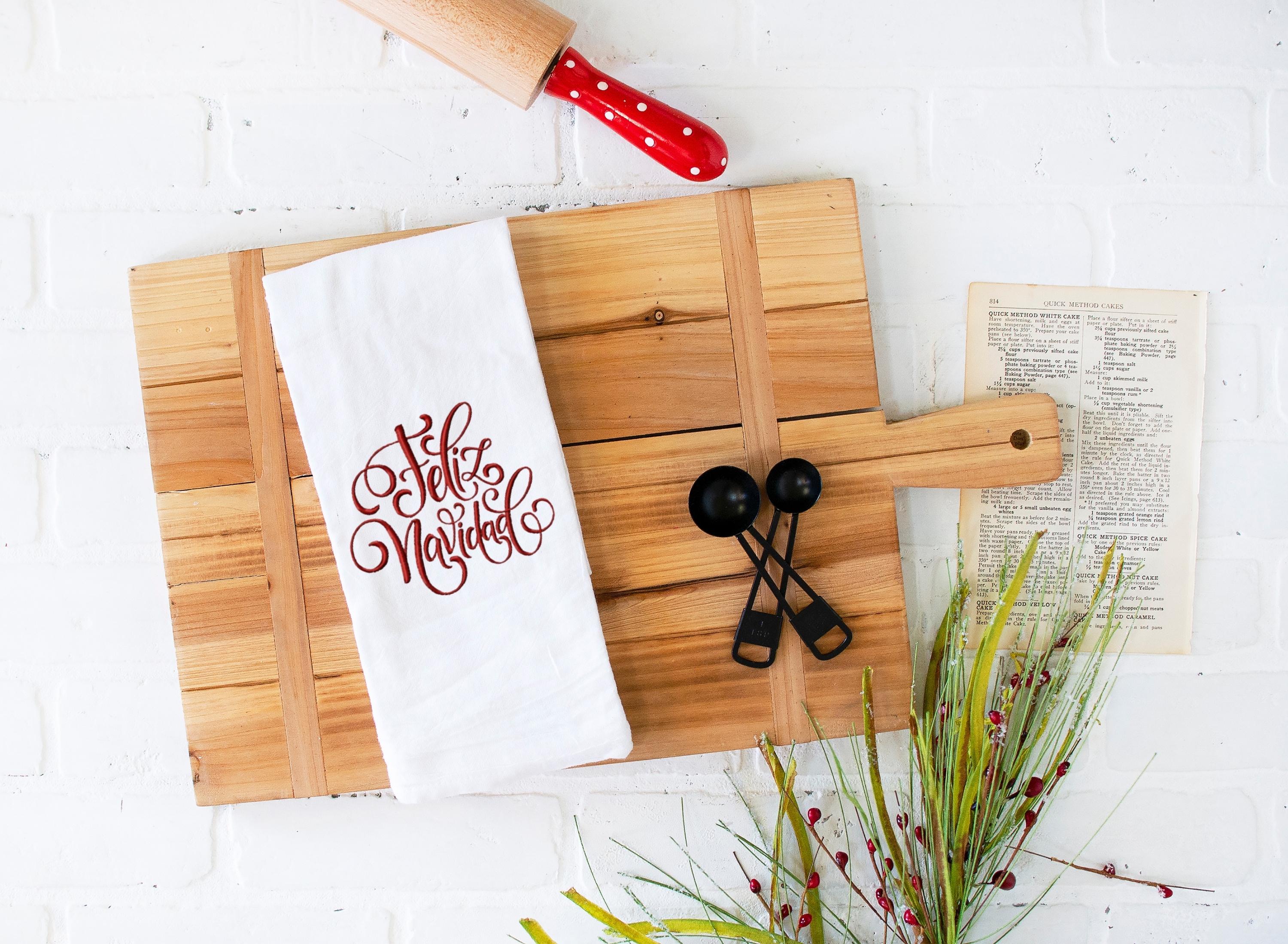 A white towel on a cutting board with Feliz Navidad embroidered in red on the towel. There is a recipe to the right and a polka dot handled rolling pin on top with Christmas greens on the bottom.