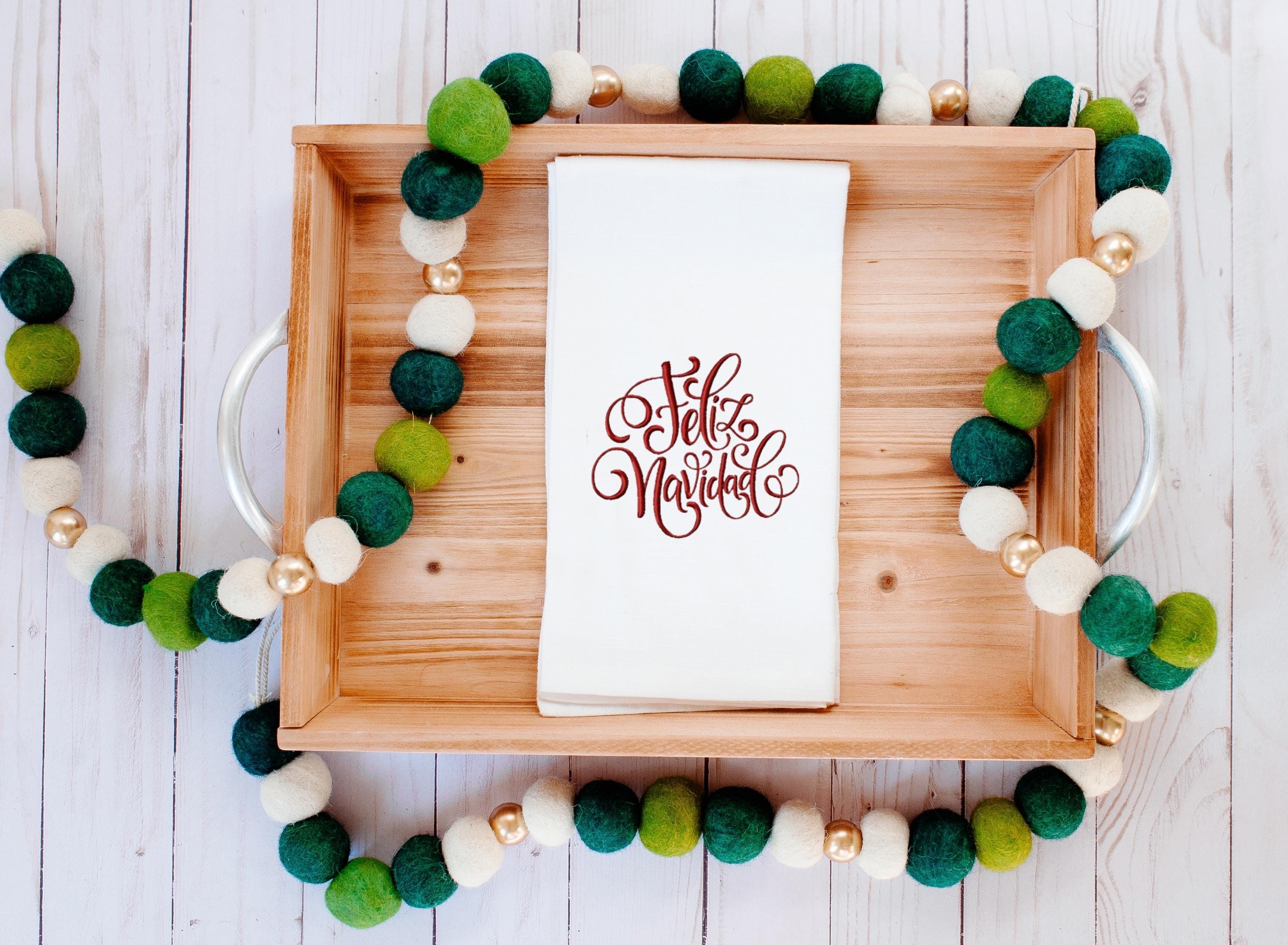 A white tea towel with Feliz Navidad embroidered in red on a wooden tray with a multi-color green and white garland on a white table.