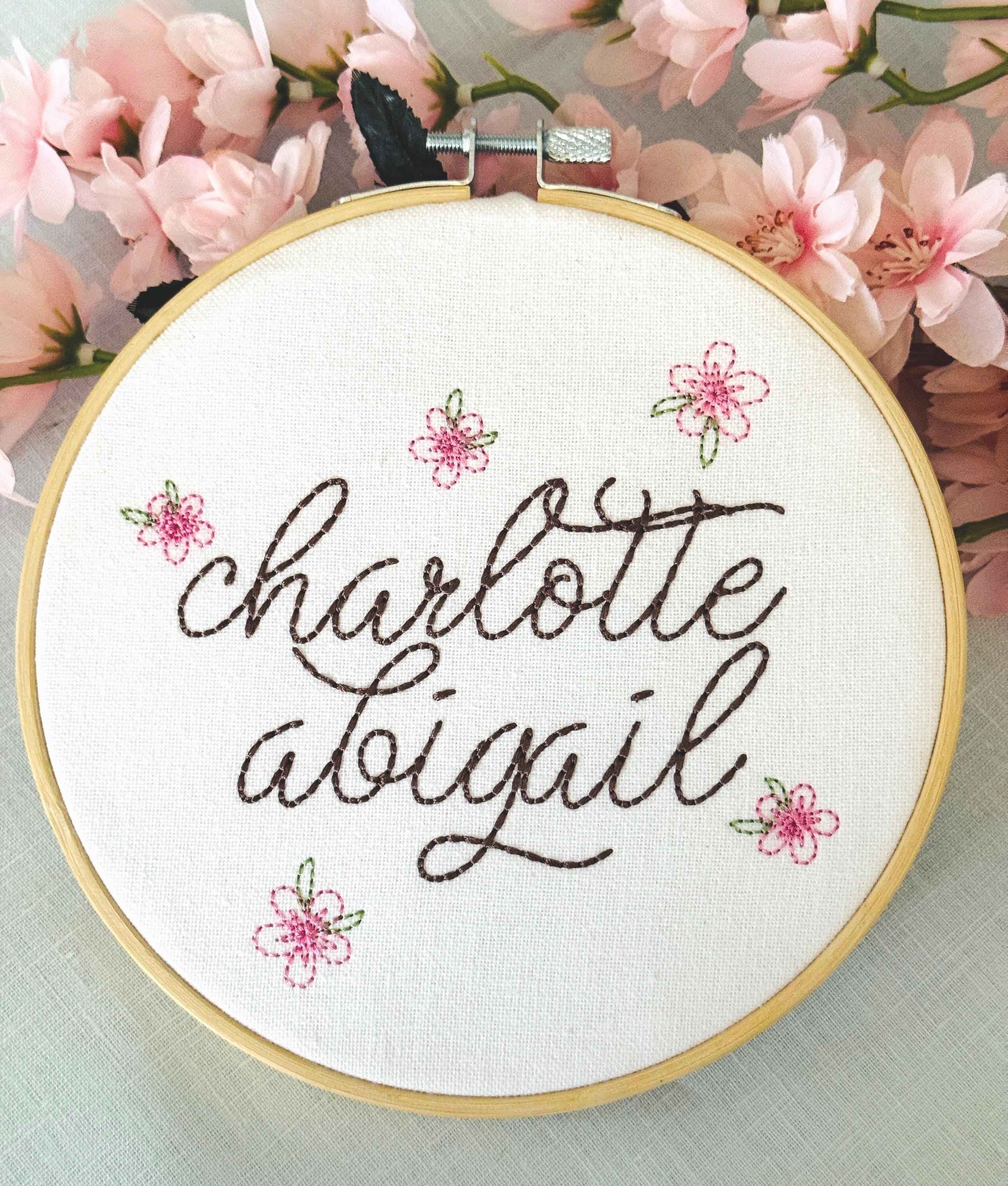 A light wood embroidery hoop art being held in one hand. The hoop has five pink embroidered flowers around the name: Charlotte Abigail in cursive. It is stitched on white cotton fabric. There are pink flower stems on top.