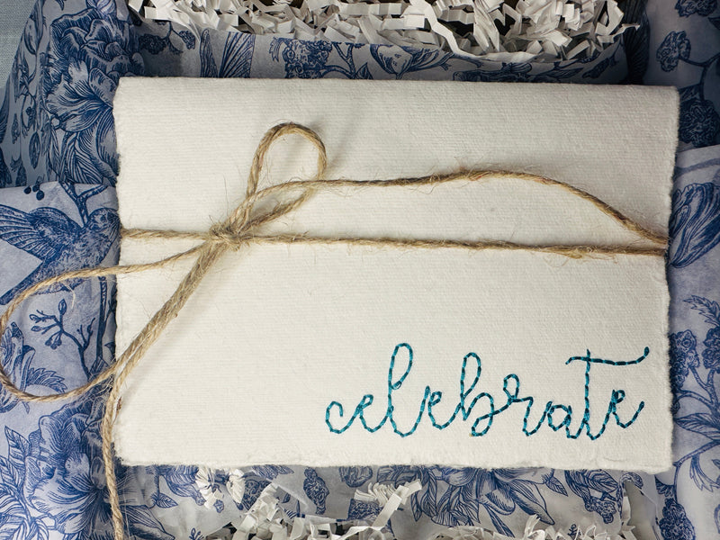 Handmade cards on deckled edge paper embroidered in teal with the word "celebrate" tied with a jute string and packaged in a box with blue and white chinoiserie tissue and white crinkle paper.