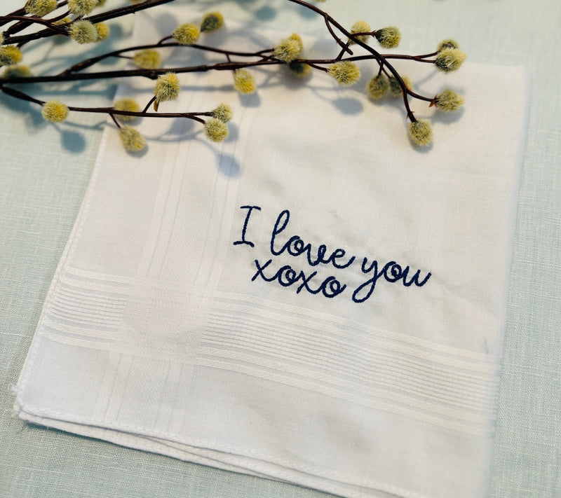 Embroidered handkerchief for groom or father of the bride. White with blue stitching. Says: I love you, xoxo. Set on a light blue tablecloth with branches on top.