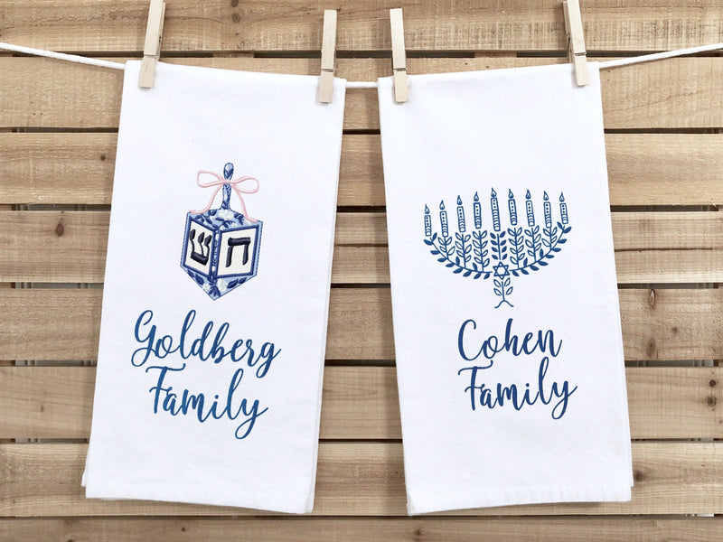 Two custom personalized Hanukkah decor embroidered towels hanging on a line stitched in blues and customized with family names. There is a dreidel towel and a menorah towel.