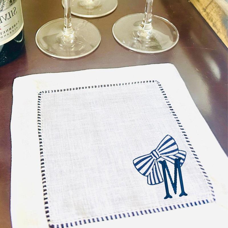 White linen cocktail napkin with a blue and white striped bow and the letter M personalized on it on the bottom right corner.