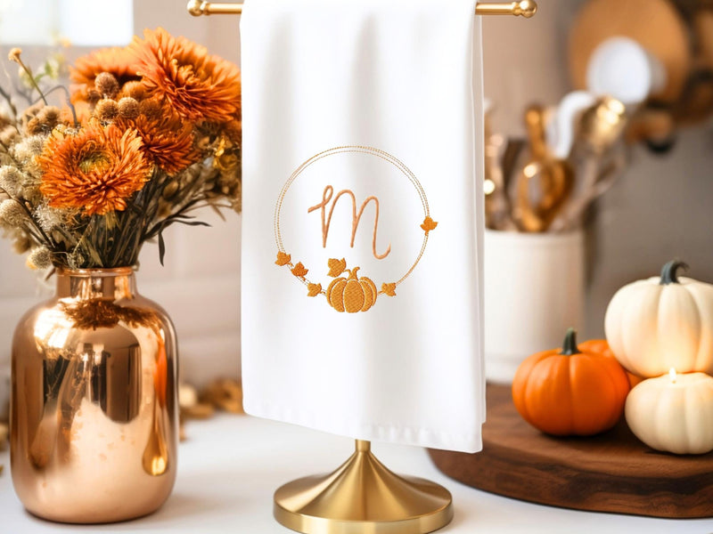 Monogrammed Pumpkin Tea Towel with an M monogram on a white towel on a countertop holder. Set on a white counter with pumpkins and a brass vase.