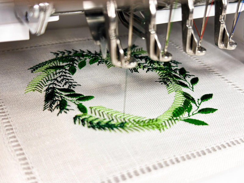 A wreath in different shades of green being embroidered onto a white linen cocktail napkin.