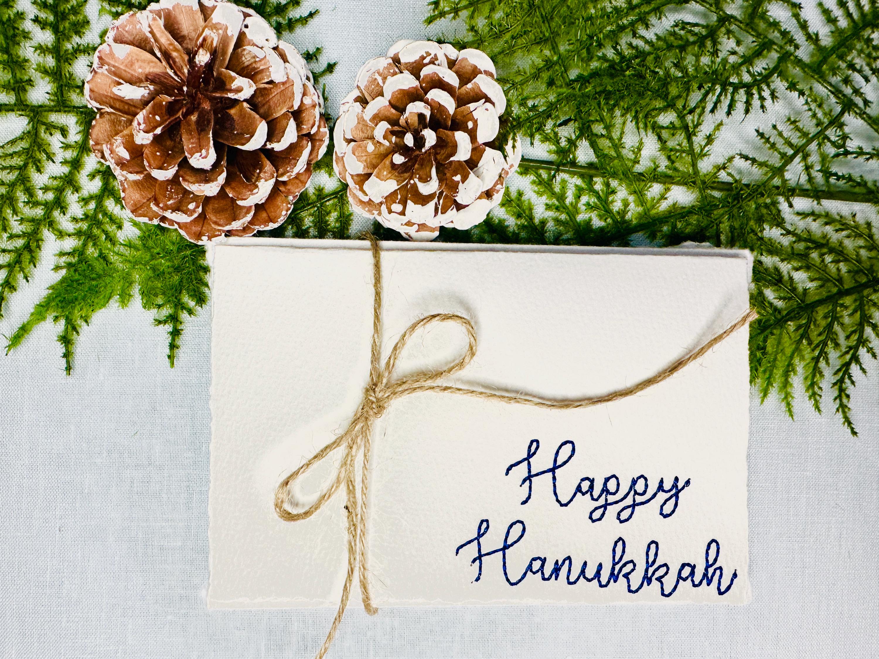 Happy Hanukkah embroidered on a 4x6 cream colored card on a table with greens and acorns