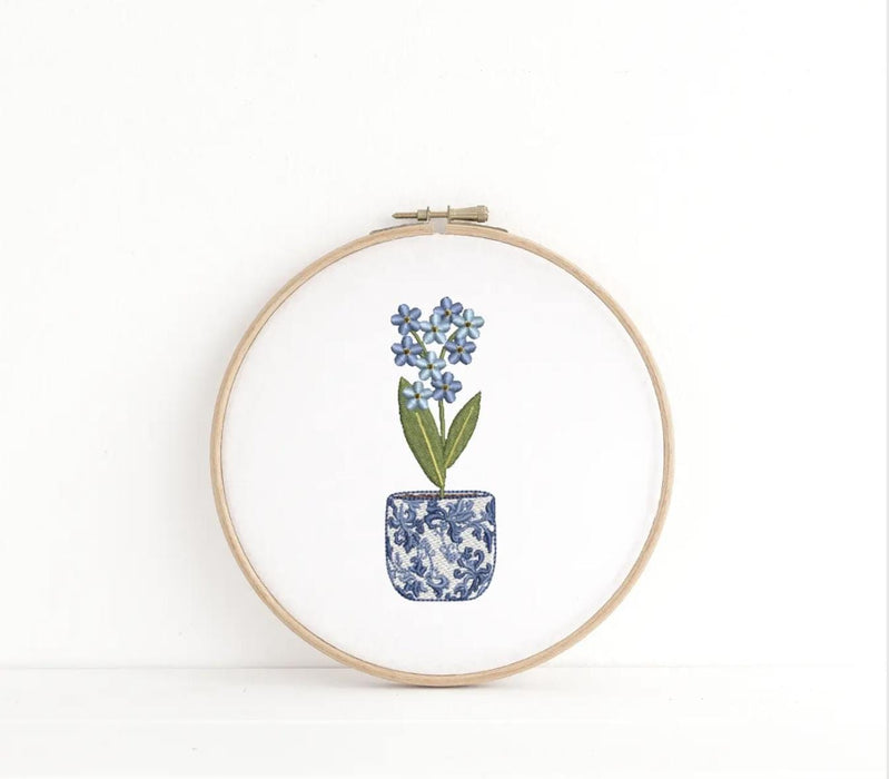 A chinoiserie pot holding a small flower with blue petals embroidered on cotton fabric and held by an embroidery hoop.