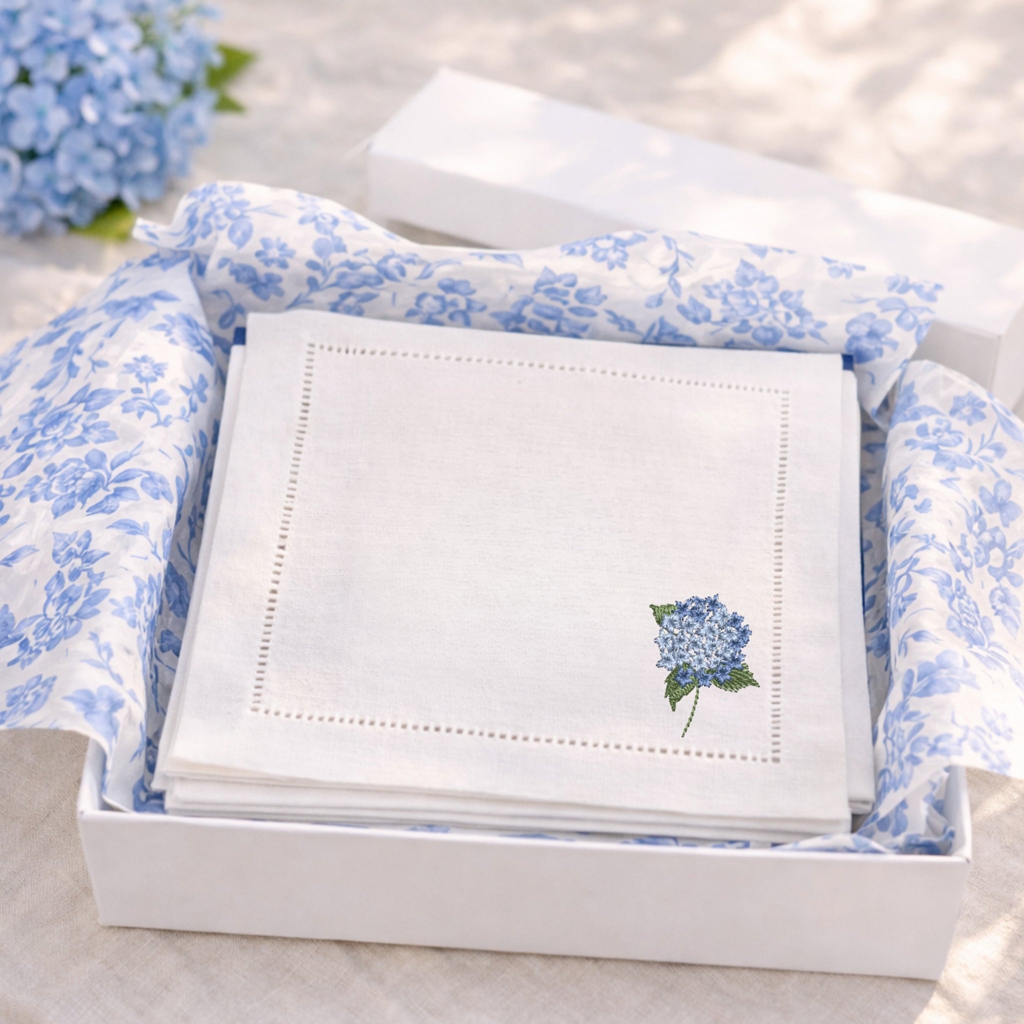 White linen cocktail napkins with an embroidered mini hydrangea in the bottom right corner in blue in a white gift box with chinoiserie blue and white tissue paper ready for gifting.