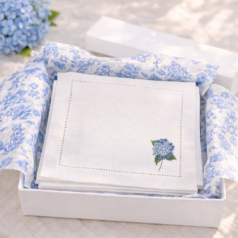 White linen cocktail napkins with an embroidered mini hydrangea in the bottom right corner in blue in a white gift box with chinoiserie blue and white tissue paper ready for gifting.