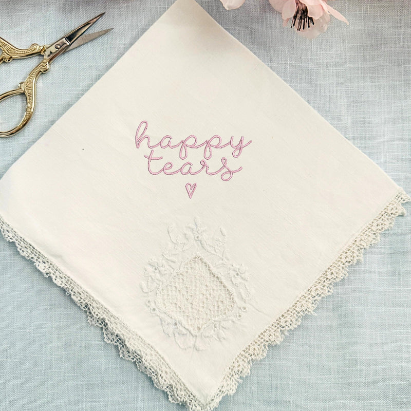 A vintage handkerchief with the words "happy tears" and a heart embroidered on it. The handkerchief is set on a light blue linen tablecloth with embroidery scissors to the top left and cherry blossoms on top.