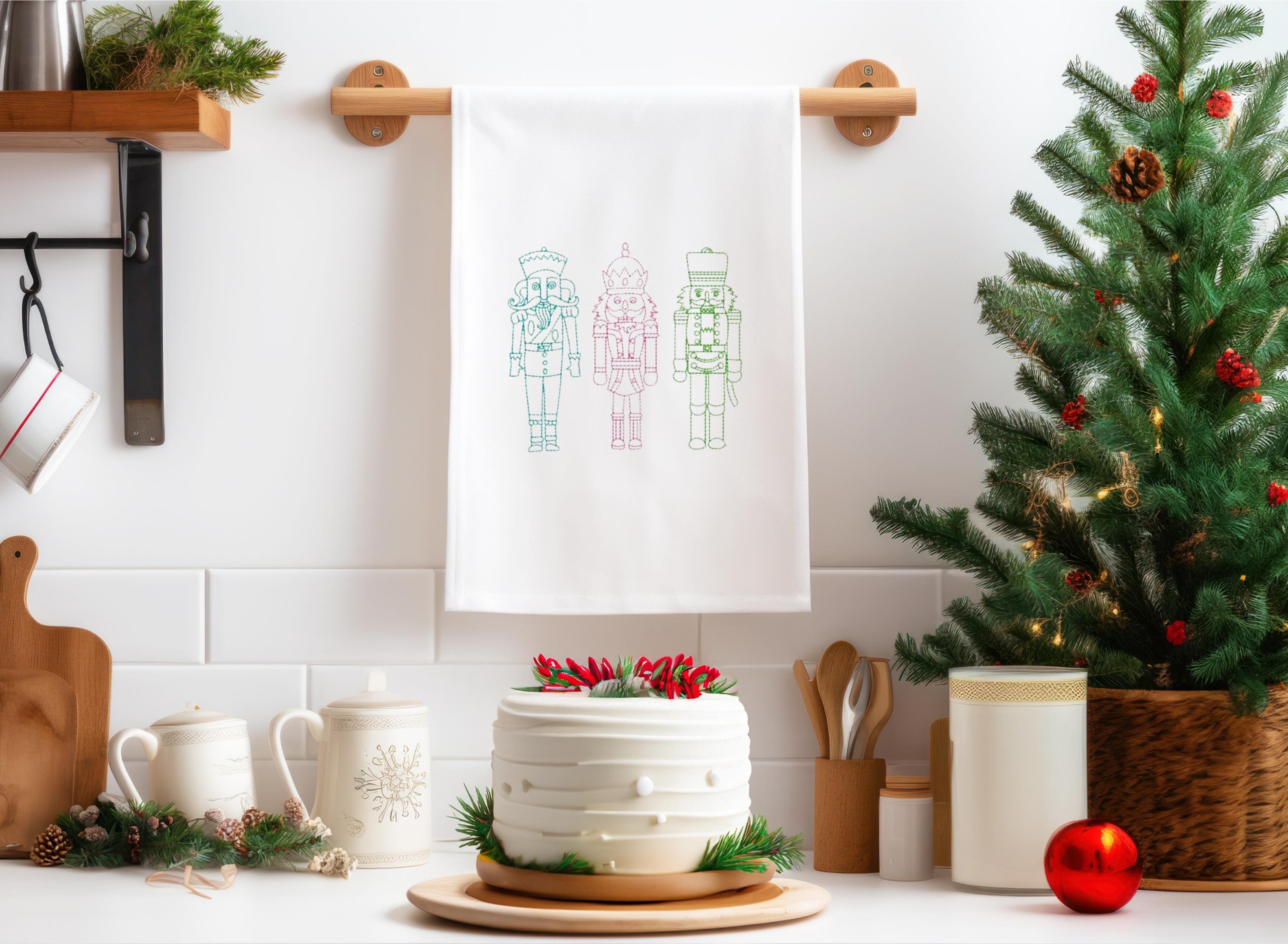 A white kitchen towel hung on the kitchen wall with three embroidered nutcrackers in pastel shades.