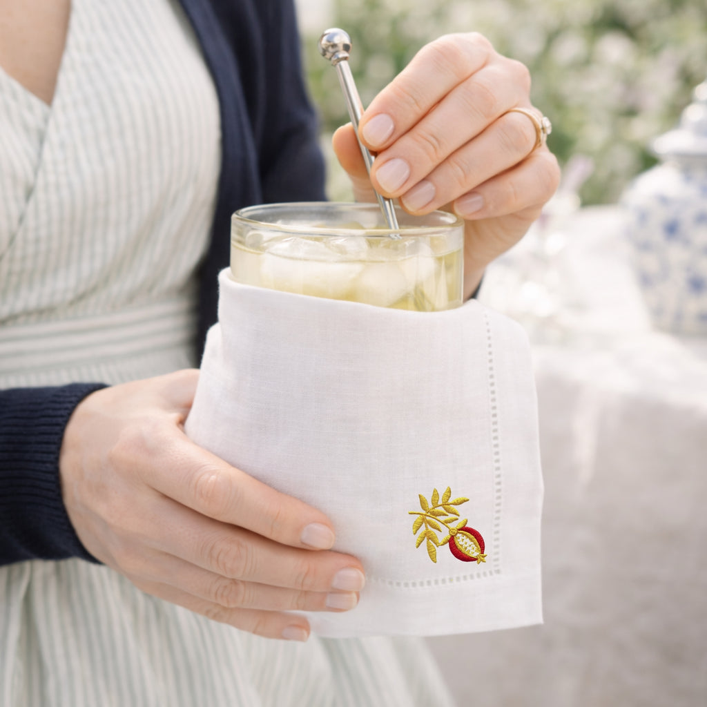 Person holding a glass of lemonade with a white napkin featuring an embroidered pomegranate outdoors.
