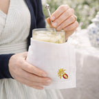 Person holding a glass of lemonade with a white napkin featuring an embroidered pomegranate outdoors.
