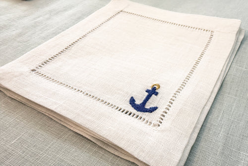 white 6" square cocktail napkin with an embroidered anchor design on a light blue linen surface