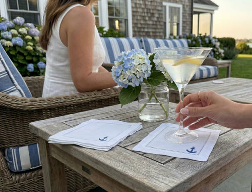 Person holding a cocktail at an outdoor table with a woman sitting on a bench, surrounded by flowers and a house in the background. The cocktail napkins are white linen with a blue embroidered anchor.