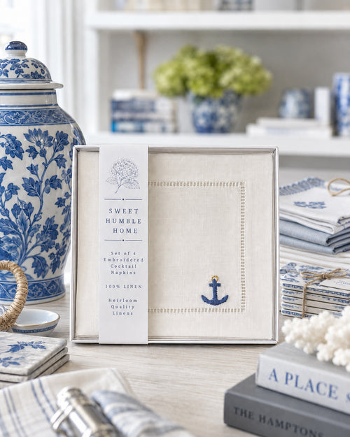 Set of 4 white linen cocktail napkins with an embroidered blue anchor with gold loop on the bottom right corner of a hemstitched napkin. On display in a styled Hampton's aesthetic