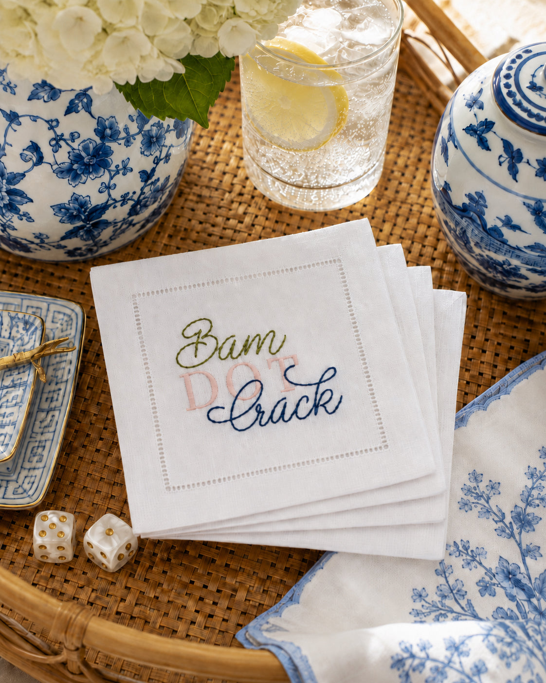 A stack of 4 white linen embroidered napkins on a woven tray, embroidered with  'Bam  DOT Crack' in green and blue script. Blue-and-white floral ceramic dishes, a glass of lemonade, mahjong dice, and flowers are arranged around it in a styled tabletop setting.