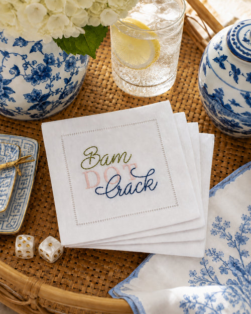 A stack of 4 white linen embroidered napkins on a woven tray, embroidered with  'Bam  DOT Crack' in green and blue script. Blue-and-white floral ceramic dishes, a glass of lemonade, mahjong dice, and flowers are arranged around it in a styled tabletop setting.