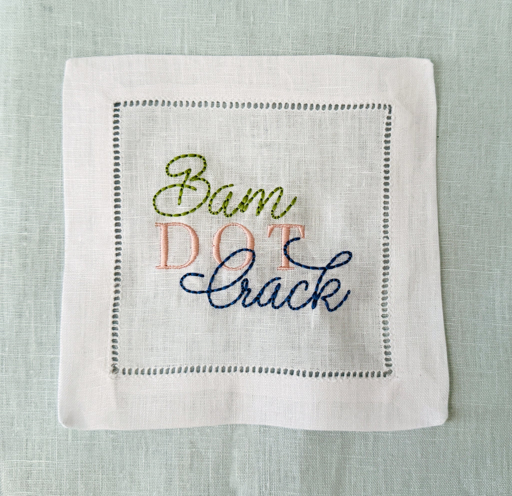 A square white linen cocktail napkin with hemstitch border stitching and embroidered text reading 'Bam Dot Crack' in olive green, pink, and navy blue thread, laid flat on a light blue linen background.