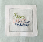 A square white linen cocktail napkin with hemstitch border stitching and embroidered text reading 'Bam Dot Crack' in olive green, pink, and navy blue thread, laid flat on a light blue linen background.