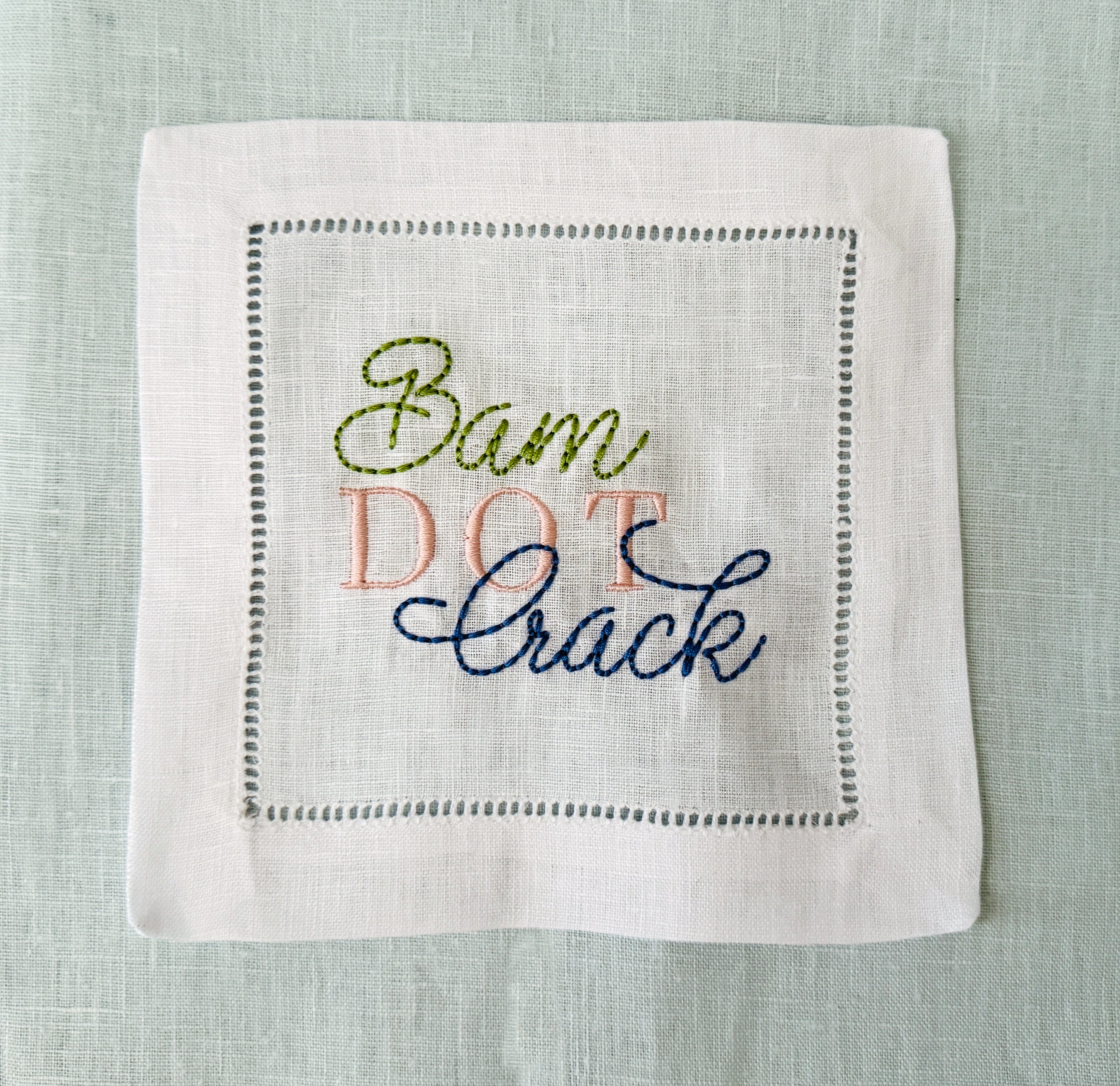 A square white linen cocktail napkin with hemstitch border stitching and embroidered text reading 'Bam Dot Crack' in olive green, pink, and navy blue thread, laid flat on a light blue linen background.