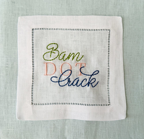 A square white linen cocktail napkin with hemstitch border stitching and embroidered text reading 'Bam Dot Crack' in olive green, pink, and navy blue thread, laid flat on a light blue linen background.