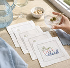 Four white woven Mahjong-themed linen coasters with embroidered designs are arranged on a light wood table in a lifestyle setting next to a window. A hand is holding a martini with olives, and other items like a blue textured pitcher, a glass of white wine, and a small bowl of nuts are visible in the background.