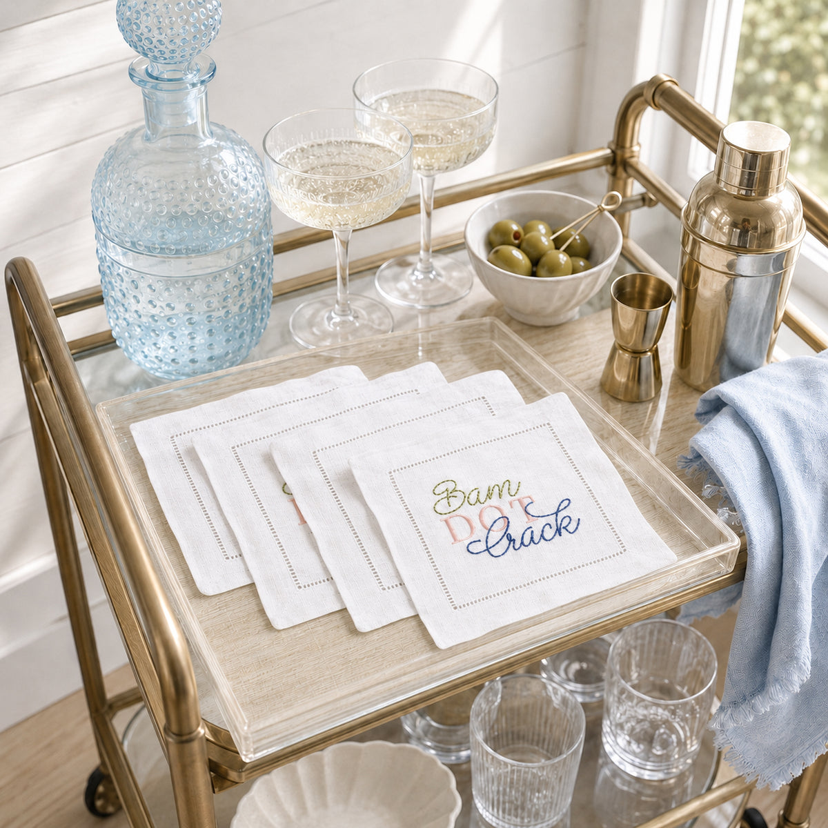 A set of white embroidered linen cocktail napkins with a 'Bam Dot Crack' design, styled on a gold bar cart alongside a blue glass decanter, coupe glasses, olives, and a shaker.