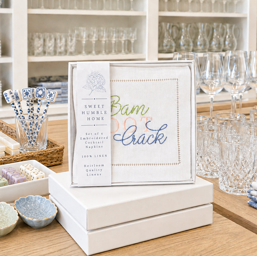 A boxed set of embroidered linen napkins displayed on stacked white boxes on a wooden table, with retail kitchen display in the background. The front napkin is seen in the packaging and shows multicolored embroidery reading 'Bam DOT Crack,' and the band on the package reads 'Sweet Humble Home' and 'Embroidered Cocktail Napkins.